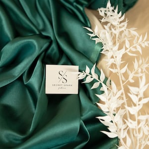 Puede incluir: Primer plano de una tela de satén verde oscuro con una tarjeta blanca con el logotipo "S/S SECRET SPARK fabrics". La tela está doblada y drapeada, creando pliegues y arrugas suaves. Una rama blanca con hojas delicadas se coloca sobre la tela.