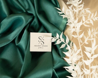 May include: A close-up of a deep green satin fabric with a white card featuring the logo "S/S SECRET SPARK fabrics". The fabric is folded and draped, creating soft folds and wrinkles. A white branch with delicate leaves is placed on the fabric.