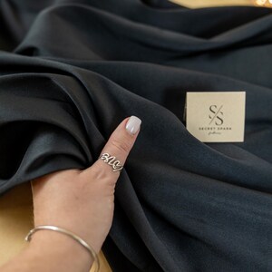 May include: A close-up of a dark gray fabric with a hand holding a silver ring. The fabric is draped over a light brown surface. A small brown box with the text "SECRET SPARK fabrics" is visible in the background.