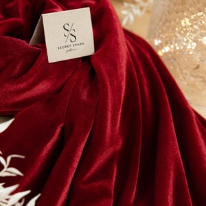 May include: A close-up of a deep red velvet fabric with soft folds. A small, square tag with the "SECRET SPARK fabrics" logo is visible. White floral accents and a blurred background add to the elegant aesthetic.