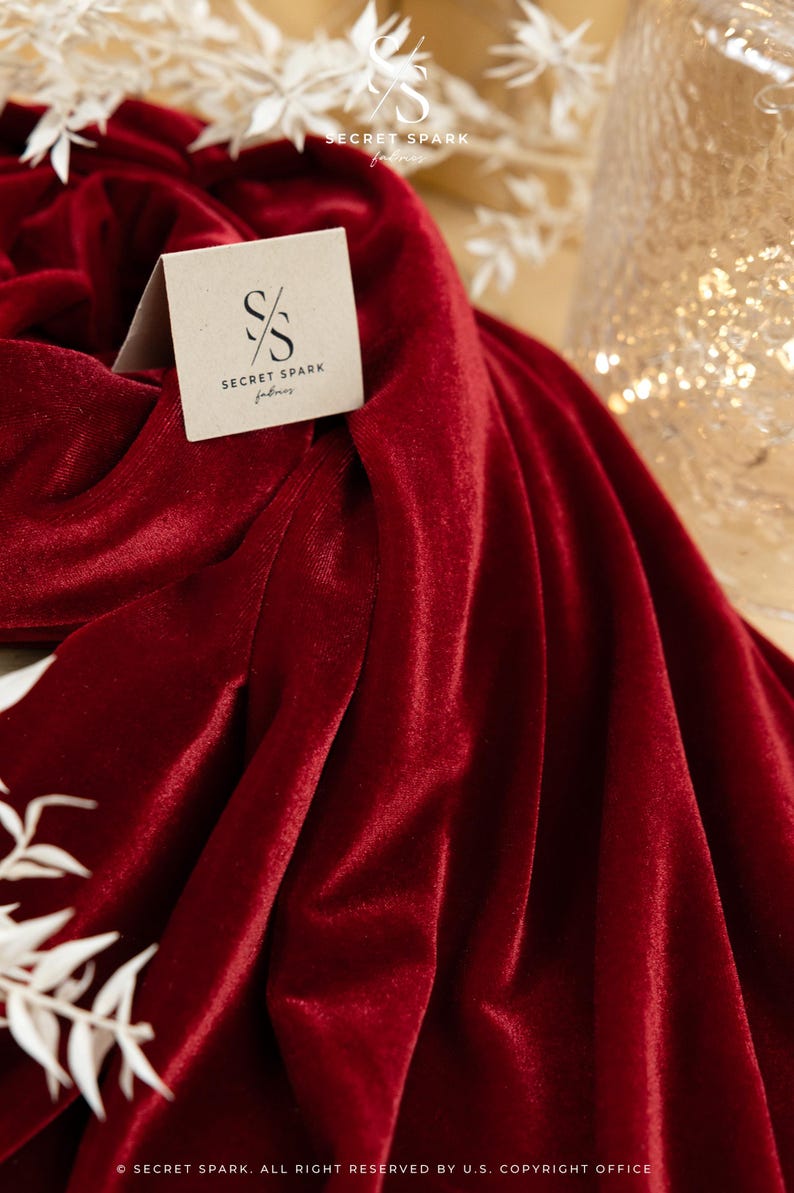 May include: A close-up of a deep red velvet fabric with soft folds. A small, square tag with the "SECRET SPARK fabrics" logo is visible. White floral accents and a blurred background add to the elegant aesthetic.