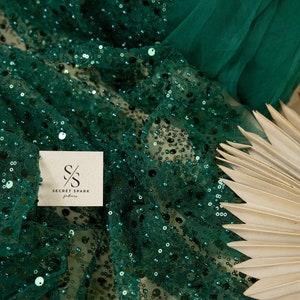 May include: Emerald green sequin fabric with a sheer mesh backing. The fabric is draped over a light brown fan. The fabric has the logo "SECRET SPARK fabrics" printed on it.