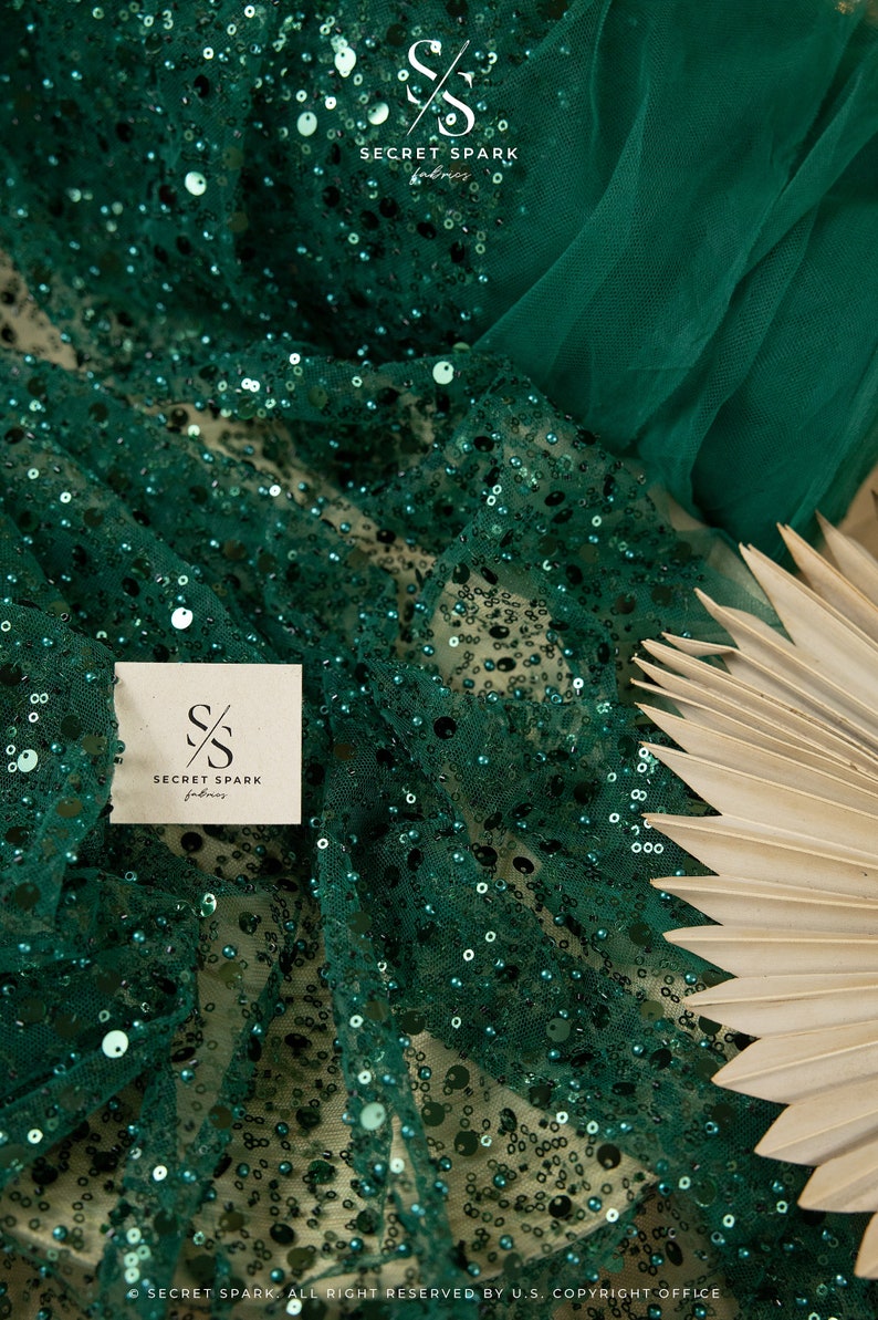 May include: Emerald green sequin fabric with a sheer mesh backing. The fabric is draped over a light brown fan. The fabric has the logo "SECRET SPARK fabrics" printed on it.