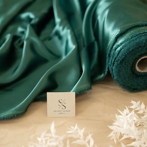 Puede incluir: Un rollo de tela de sat&eacute;n verde azulado con una etiqueta blanca que dice "SECRET SPARK fabrics".