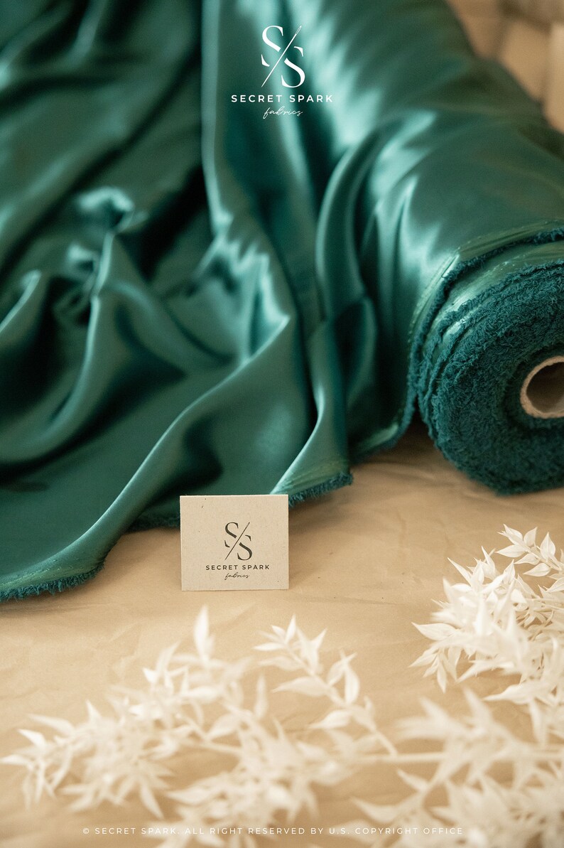 Puede incluir: Un rollo de tela de sat&eacute;n verde azulado con una etiqueta blanca que dice "SECRET SPARK fabrics".