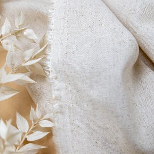 May include: A piece of off-white, textured fabric with a visible weave and a frayed, raw edge. The material features small, darker flecks throughout its surface. A logo with the text SECRET SPARK is visible at the top of the fabric.