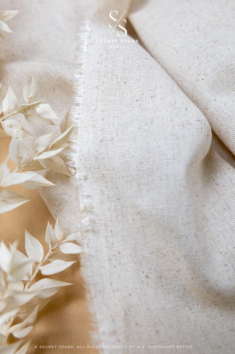 May include: A piece of off-white, textured fabric with a visible weave and a frayed, raw edge. The material features small, darker flecks throughout its surface. A logo with the text SECRET SPARK is visible at the top of the fabric.