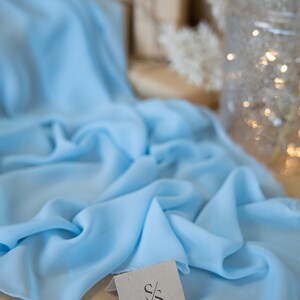 May include: Light blue fabric draped across a surface. The fabric has a soft, flowing appearance. A small tag with the "Secret Spark Fabrics" logo is visible. The background includes a blurred decorative vase and dried flowers.