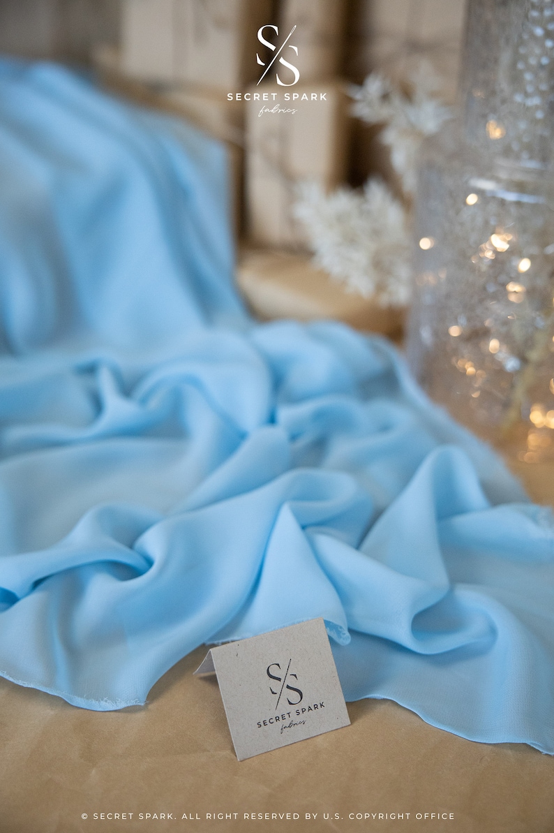 May include: Light blue fabric draped across a surface. The fabric has a soft, flowing appearance. A small tag with the "Secret Spark Fabrics" logo is visible. The background includes a blurred decorative vase and dried flowers.