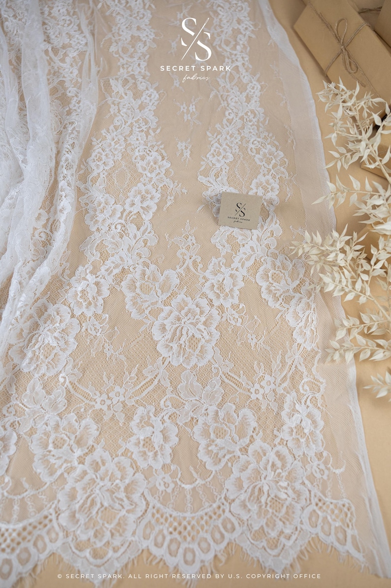 May include: White floral lace fabric with a delicate pattern. The lace is displayed on a beige background, with the text "SECRET SPARK fabrics" visible. The fabric is suitable for bridal wear and other craft projects.