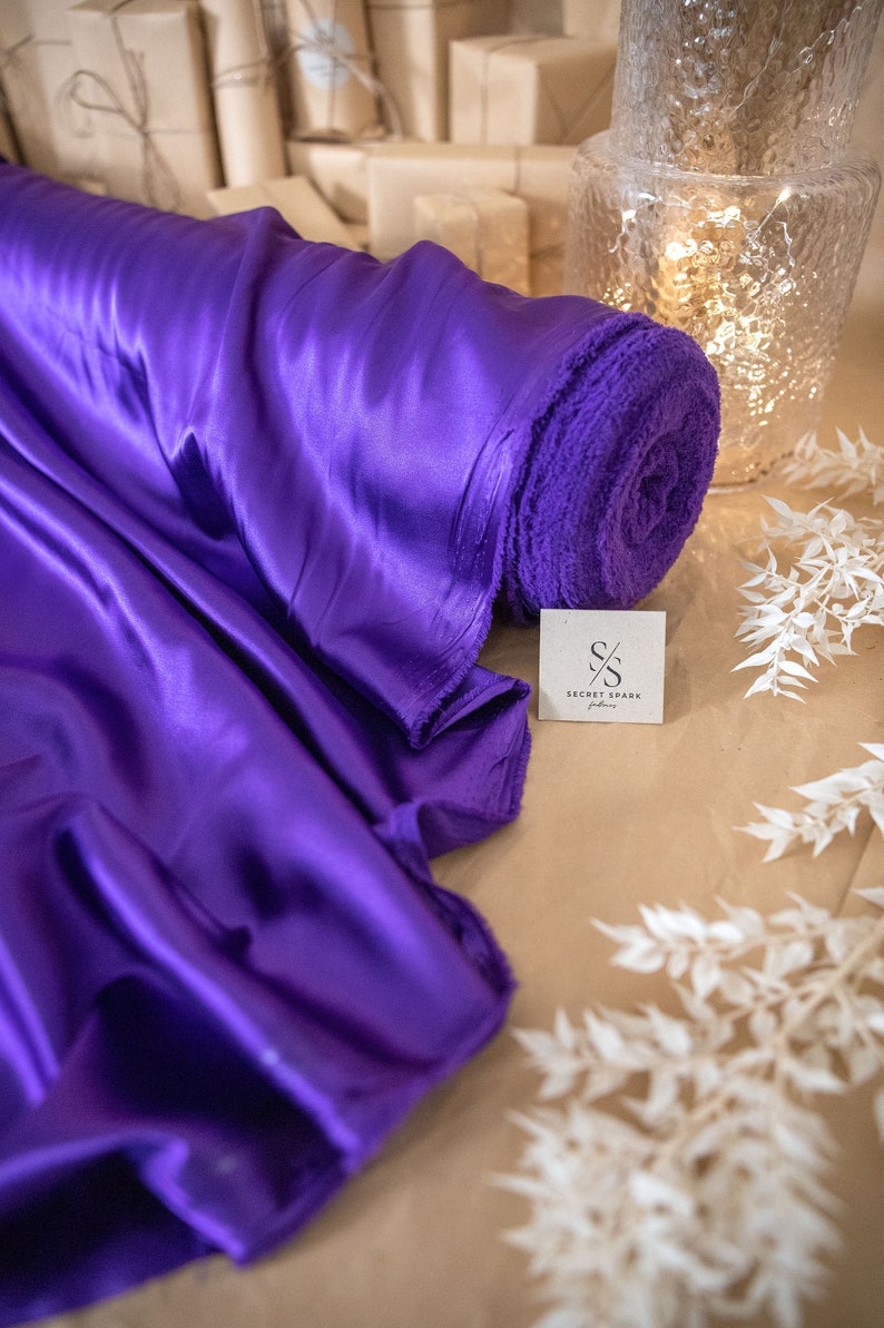 Pu&ograve; includere: Un rotolo di tessuto di raso viola con un'etichetta che dice "Secret Spark Fabrics".