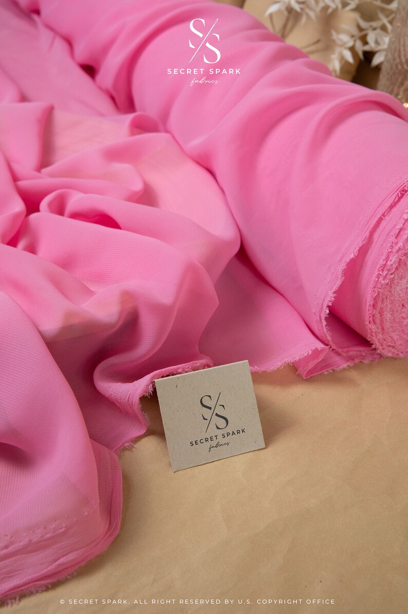 May include: Pink chiffon fabric rolled up on a brown surface. The fabric is soft and sheer, with a subtle sheen. The fabric is labeled "SECRET SPARK fabrics".