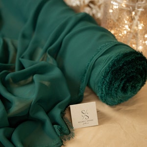 May include: A roll of emerald green chiffon fabric with a fringed edge. The fabric is rolled up and sitting on a brown surface. The Secret Spark Fabrics logo is visible on a small card in the foreground.