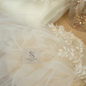 May include: A roll of ivory tulle fabric with a small brown card that says "SECRET SPARK fabrics" on it. The fabric is draped over a wooden surface and there are white dried flowers on top of the fabric.