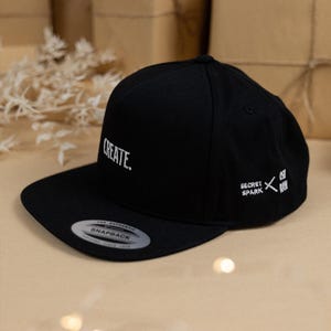 May include: Black snapback hat with the word "CREATE" in white script. The hat also features the "SECRET SPARK" logo and text. The hat has a flat brim and a snap closure.