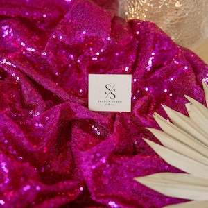 May include: A close-up of a bright pink sequined fabric with a small white square label that says "SECRET SPARK fabrics".