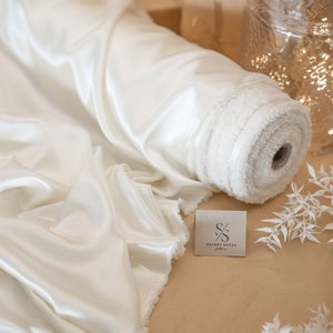 May include: A roll of white satin fabric with a frayed edge. The fabric is rolled up and tied with a white ribbon. The fabric is on a brown surface with a white tag that says "SECRET SPARK fabrics".
