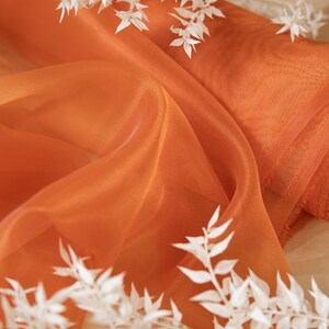 Könnte beinhalten: Orangener, durchsichtiger Stoff mit einem Verlaufseffekt, drapiert über weiße, getrocknete Zweige. Der Stoff ist durchscheinend und hat eine weiche, fließende Textur. © SECRET SPARK. ALL RIGHT RESERVED BY U.S. COPYRIGHT OFFICE