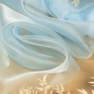 May include: A light blue sheer fabric with a subtle shimmer. The fabric is draped and folded, creating soft folds and curves. The fabric is on a brown surface with dried white flowers.