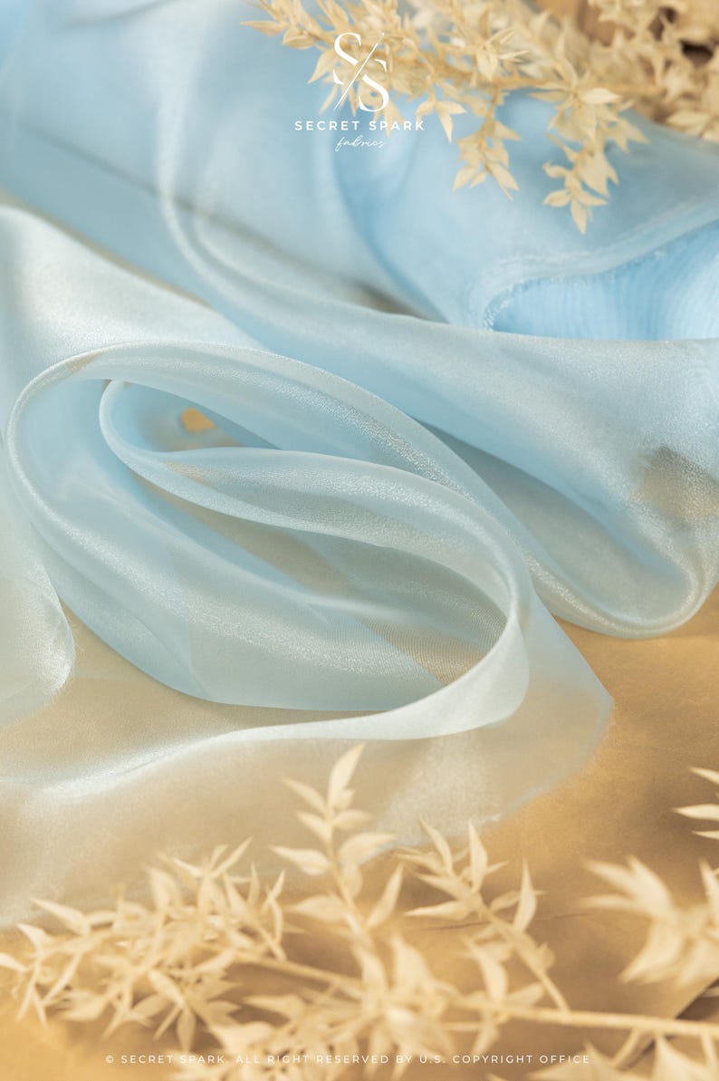May include: A light blue sheer fabric with a subtle shimmer. The fabric is draped and folded, creating soft folds and curves. The fabric is on a brown surface with dried white flowers.