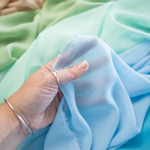 May include: A hand holding a swatch of light blue fabric, with a gradient of colors in the background. The fabric is sheer and has a delicate texture. The image includes the text "SECRET SPARK fabrics."