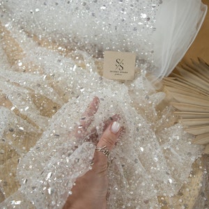 Könnte beinhalten: Weißer Tüllstoff mit silbernen Pailletten und kleinen weißen Perlen. Der Stoff ist über eine Holzoberfläche drapiert. Der Text "SECRET SPARK" ist auf einem Etikett sichtbar.