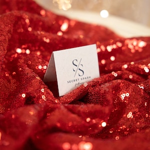 May include: A white card with the text "SECRET SPARK" and a stylized "S" logo on a red sequined fabric background.