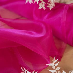 May include: A close-up of a bright pink sheer fabric, draped and folded. The fabric is translucent and has a soft, flowing texture. The fabric is accented with white dried branches.