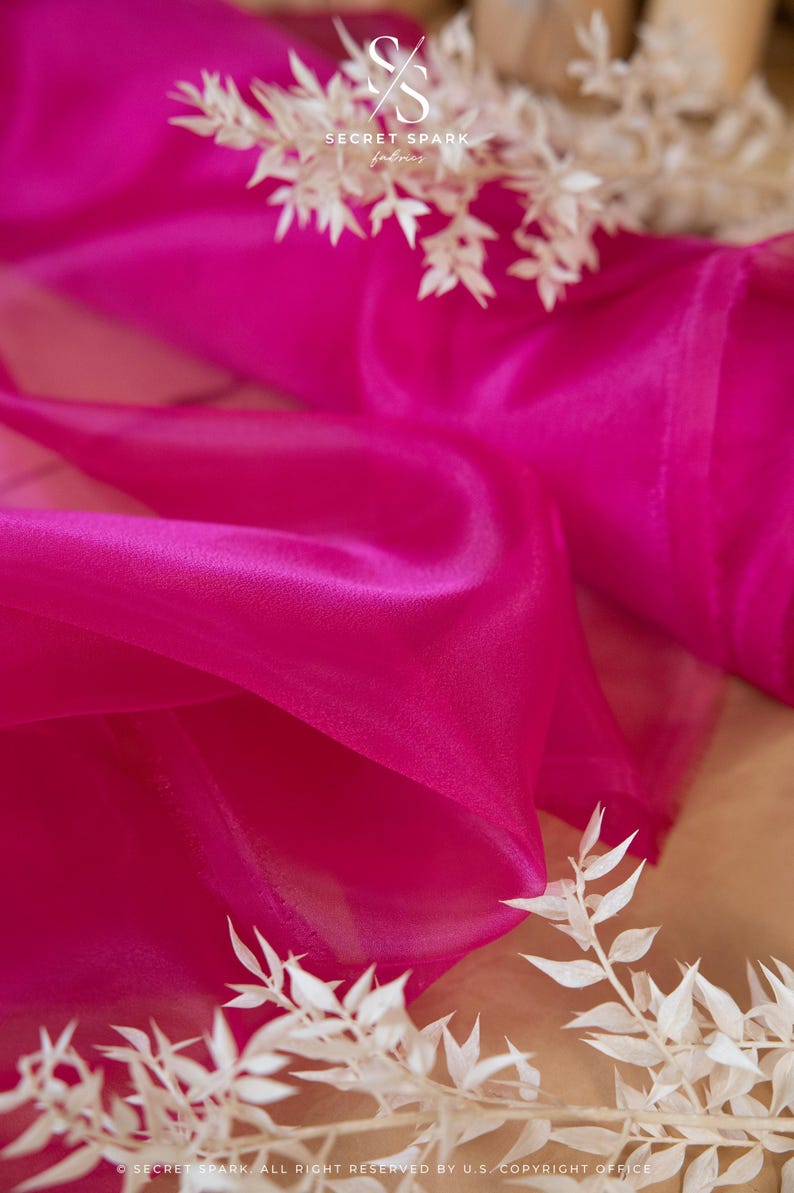 May include: A close-up of a bright pink sheer fabric, draped and folded. The fabric is translucent and has a soft, flowing texture. The fabric is accented with white dried branches.