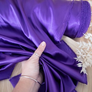 Pu&ograve; includere: Un rotolo di tessuto di raso viola. Il tessuto &egrave; liscio e lucido, ed &egrave; piegato a met&agrave;. Il tessuto &egrave; tenuto da una mano che indossa un anello d'argento.