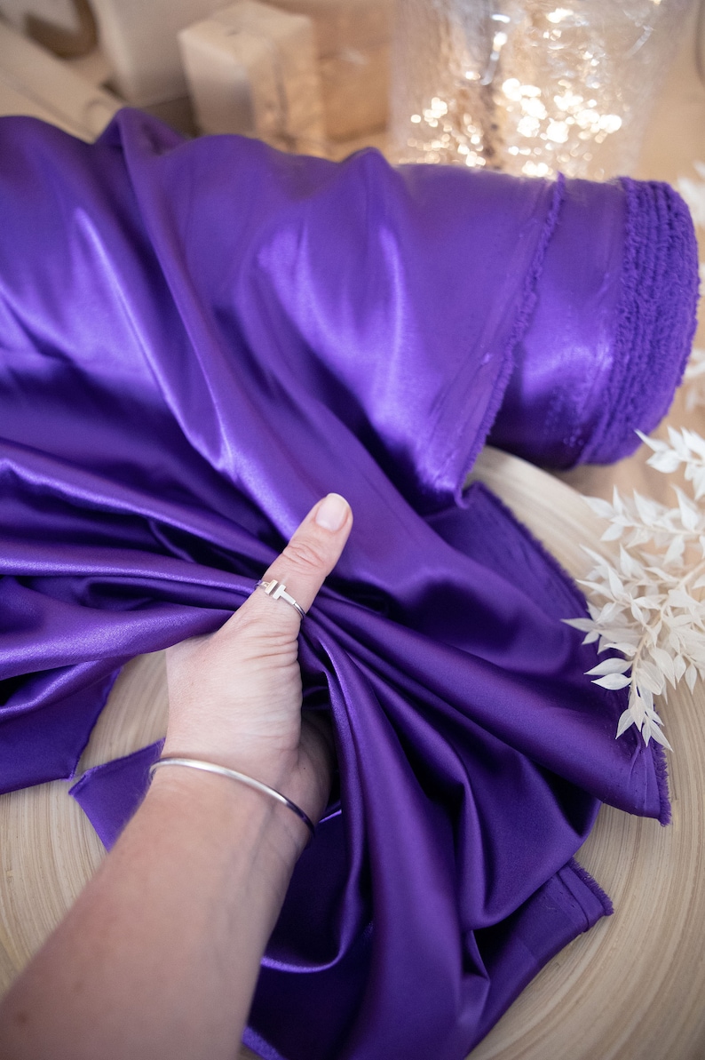 Pu&ograve; includere: Un rotolo di tessuto di raso viola. Il tessuto &egrave; liscio e lucido, ed &egrave; piegato a met&agrave;. Il tessuto &egrave; tenuto da una mano che indossa un anello d'argento.
