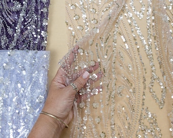 May include: Close-up of several fabrics with intricate sequin and bead embellishments. One fabric is a light beige with a floral pattern, another is a deep purple, and a third is a light blue. The fabrics are ideal for creating elegant apparel.