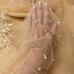 Könnte beinhalten: Beigefarbener Tüllstoff mit verstreuten Perlenbesatz. Der Stoff ist über eine Hand gelegt und zeigt die Textur und den Glanz des Materials. Der Text "SECRET SPARK fabrics" ist auf einem braunen Etikett sichtbar.