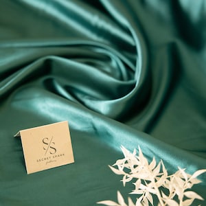Puede incluir: Una peque&ntilde;a tarjeta marr&oacute;n con el texto "SECRET SPARK" y las iniciales "SS" en una fuente estilizada. La tarjeta est&aacute; sobre una tela de sat&eacute;n turquesa con una rama blanca de hojas.