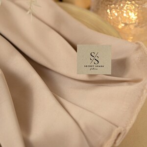May include: A close-up of a light beige fabric with a brown square label that says "SECRET SPARK fabrics".
