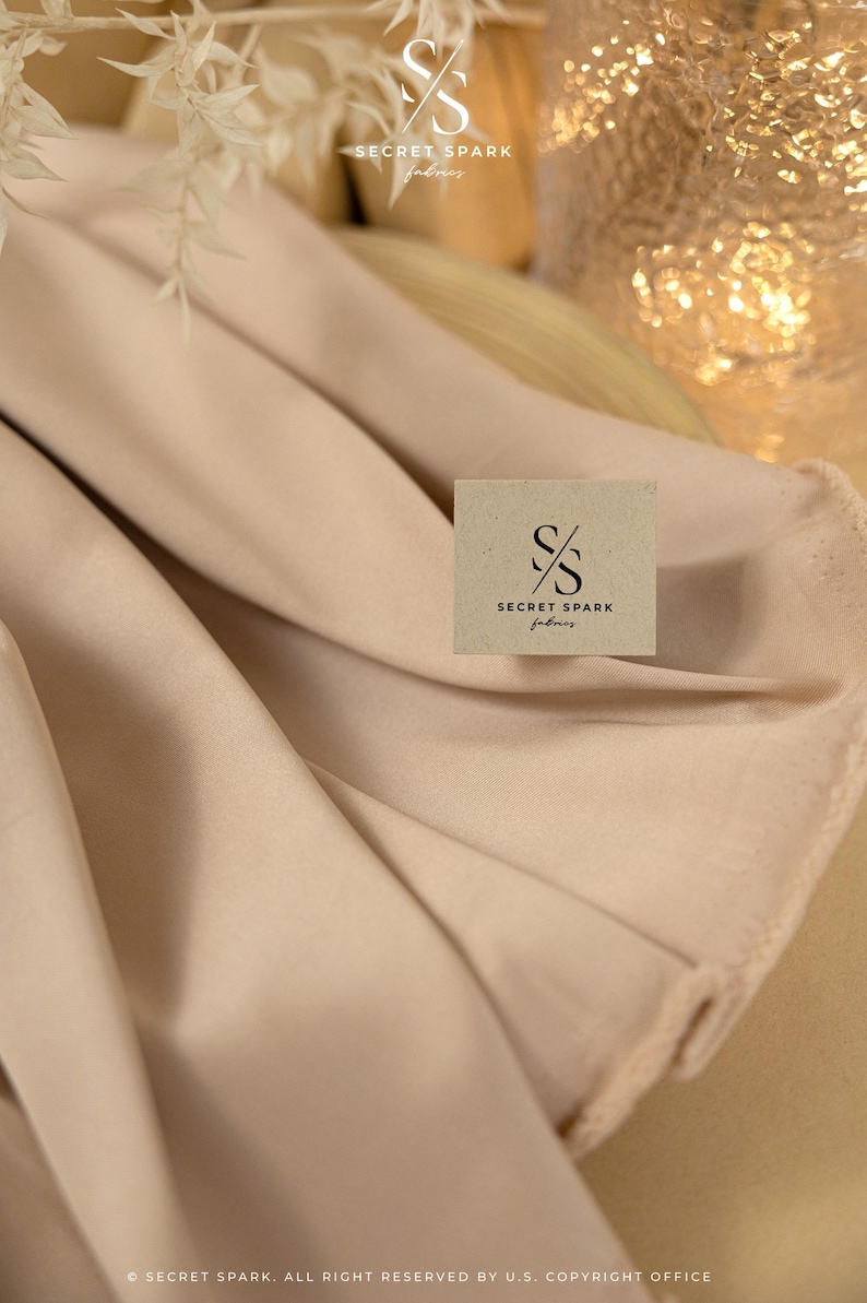 May include: A close-up of a light beige fabric with a brown square label that says "SECRET SPARK fabrics".