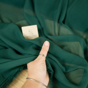 May include: A close-up of a deep green fabric with a fringed edge. The fabric is draped over a wooden surface and a hand is holding a corner of the fabric. The fabric is labeled "Secret Spark Fabrics".
