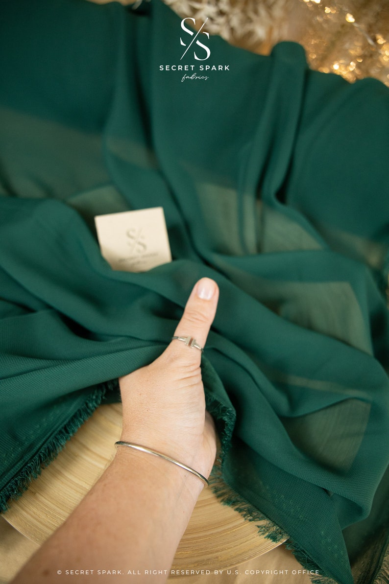 May include: A close-up of a deep green fabric with a fringed edge. The fabric is draped over a wooden surface and a hand is holding a corner of the fabric. The fabric is labeled "Secret Spark Fabrics".