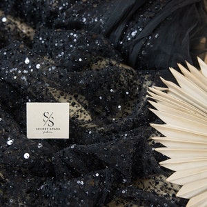 May include: Black sequined fabric with a sheer mesh base. The fabric is folded and draped over a light brown surface. A small white tag with the text "SECRET SPARK fabrics" is visible in the image.