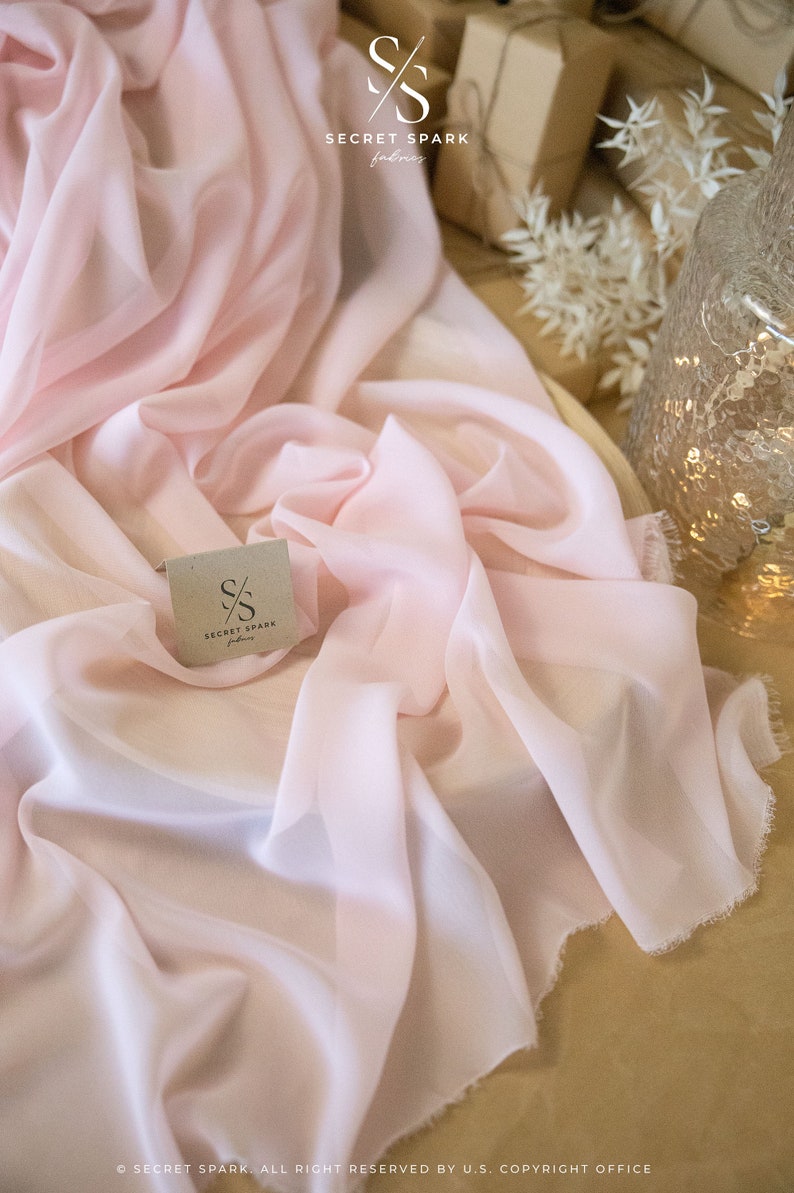 K&ouml;nnte beinhalten: Ein blasses rosa, durchsichtiges Gewebe mit ausgefransten Kanten. Der Stoff ist gefaltet und auf einer Holzoberfl&auml;che drapiert. Ein kleines wei&szlig;es Etikett mit dem Text "SECRET SPARK fabrics" ist auf dem Stoff sichtbar.