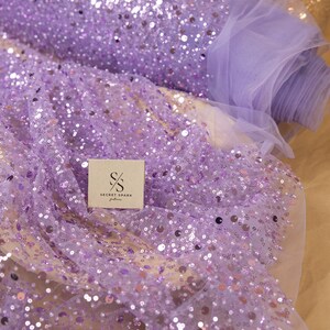 May include: Purple sequin fabric with a sheer tulle backing. The fabric is covered in small, round sequins that are randomly scattered across the surface. The fabric is rolled up and the label "SECRET SPARK fabrics" is visible.