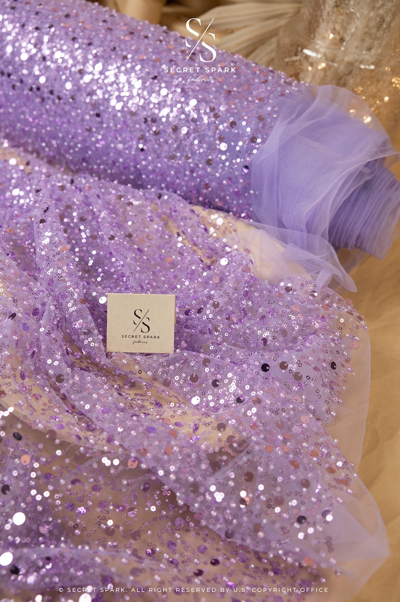 May include: Purple sequin fabric with a sheer tulle backing. The fabric is covered in small, round sequins that are randomly scattered across the surface. The fabric is rolled up and the label "SECRET SPARK fabrics" is visible.