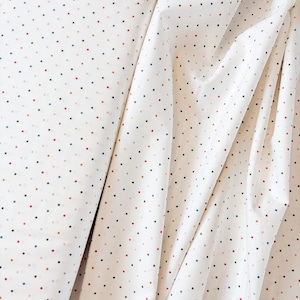 May include: White fabric with a repeating pattern of small, evenly spaced dots in various colors, including red, blue, and orange. The fabric is draped, showing folds and texture, suggesting it's a soft material.