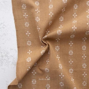May include: Close-up of a tan fabric with a repeating pattern of white floral and cross-shaped designs arranged in vertical stripes. The fabric is folded, creating shadows and texture. The word "FABLEISM" is printed in white at the bottom.
