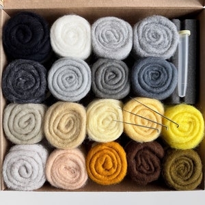 Op de afbeelding: Een set van 20 wolviltrollen in verschillende tinten grijs, beige, geel, oranje, bruin en groen, gerangschikt in een doos met drie vilten naalden en een klein buisje zeep.