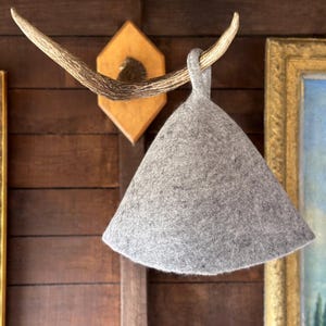 Może przedstawiać: Szary filcowy kapelusz do sauny wisi na uchwycie z poroża jelenia na drewnianej ścianie. Kapelusz ma kształt stożka z pętlą u góry. Po prawej stronie widoczny jest obraz w ramie.
