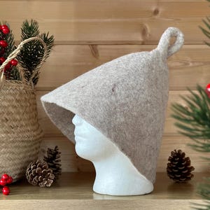 May include: A beige wool hat with a pointed crown and a loop on top. The hat is sitting on a wooden surface with pine cones and greenery.