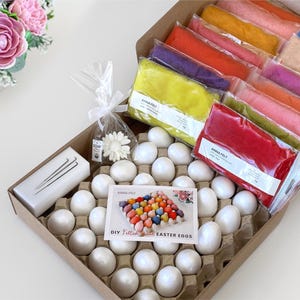 Peut inclure: Un kit de bricolage avec une variété de feuilles de feutre colorées, des œufs blancs et une carte indiquant "DIY Felted Wool Easter Eggs". Le kit comprend une boîte d'œufs, du feutre de différentes couleurs et un petit sac avec une fleur.