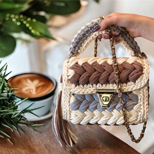 Op de afbeelding: Een gehaakte handtas in de kleuren bruin, wit en lichtblauw. De tas heeft een gouden kettingband en een klein gouden sluiting. De tas ligt op een houten tafel met een kop koffie op de achtergrond.