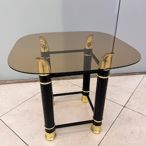 Può includere: Un tavolino quadrato con piano in vetro fumé. Il tavolo ha gambe nere con dettagli color oro nella parte superiore e inferiore. Il tavolo misura circa 61 cm di larghezza e 61 cm di lunghezza.